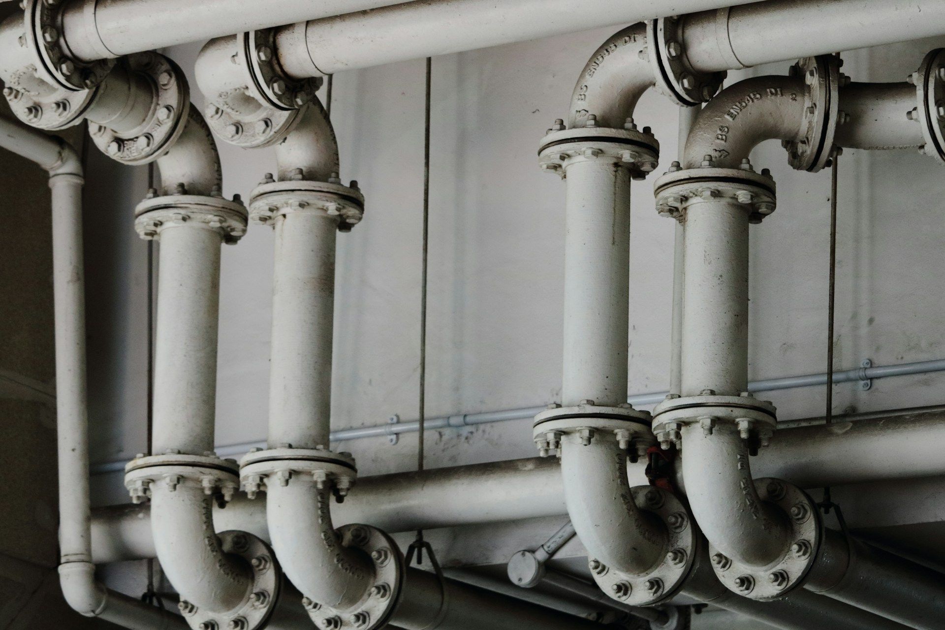 Industrial white piping system with multiple 90-degree elbows and flanged joints mounted against a grey wall.