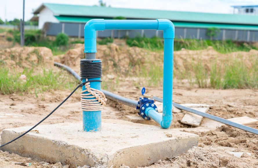 A blue PVC pipe assembly connected to a ground well, situated on a concrete slab in an open, dirt-filled field.