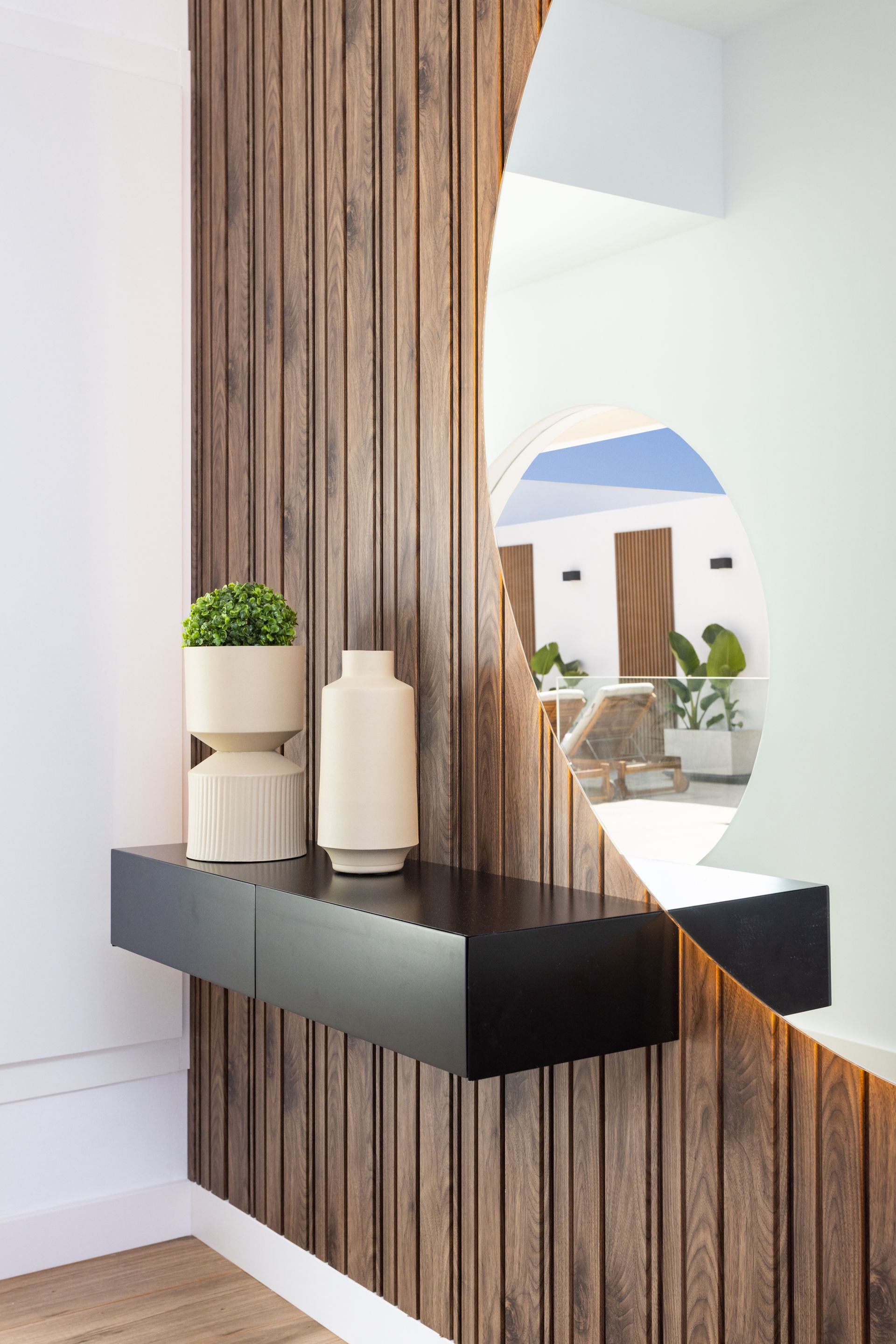 Modern entryway with vertical wood slats, black console shelf, and a round mirror reflecting a bright room.