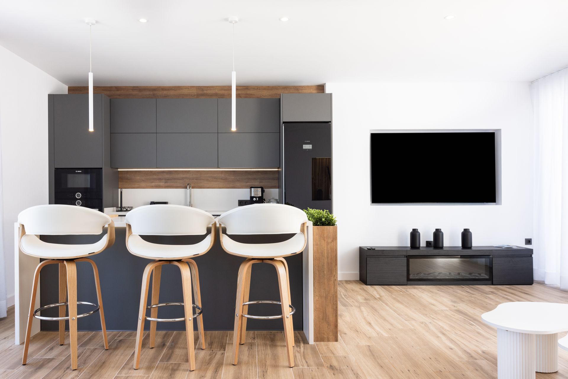 Cocina y sala de estar modernas de concepto abierto con taburetes de bar, suelos de madera y un televisor montado en la pared.
