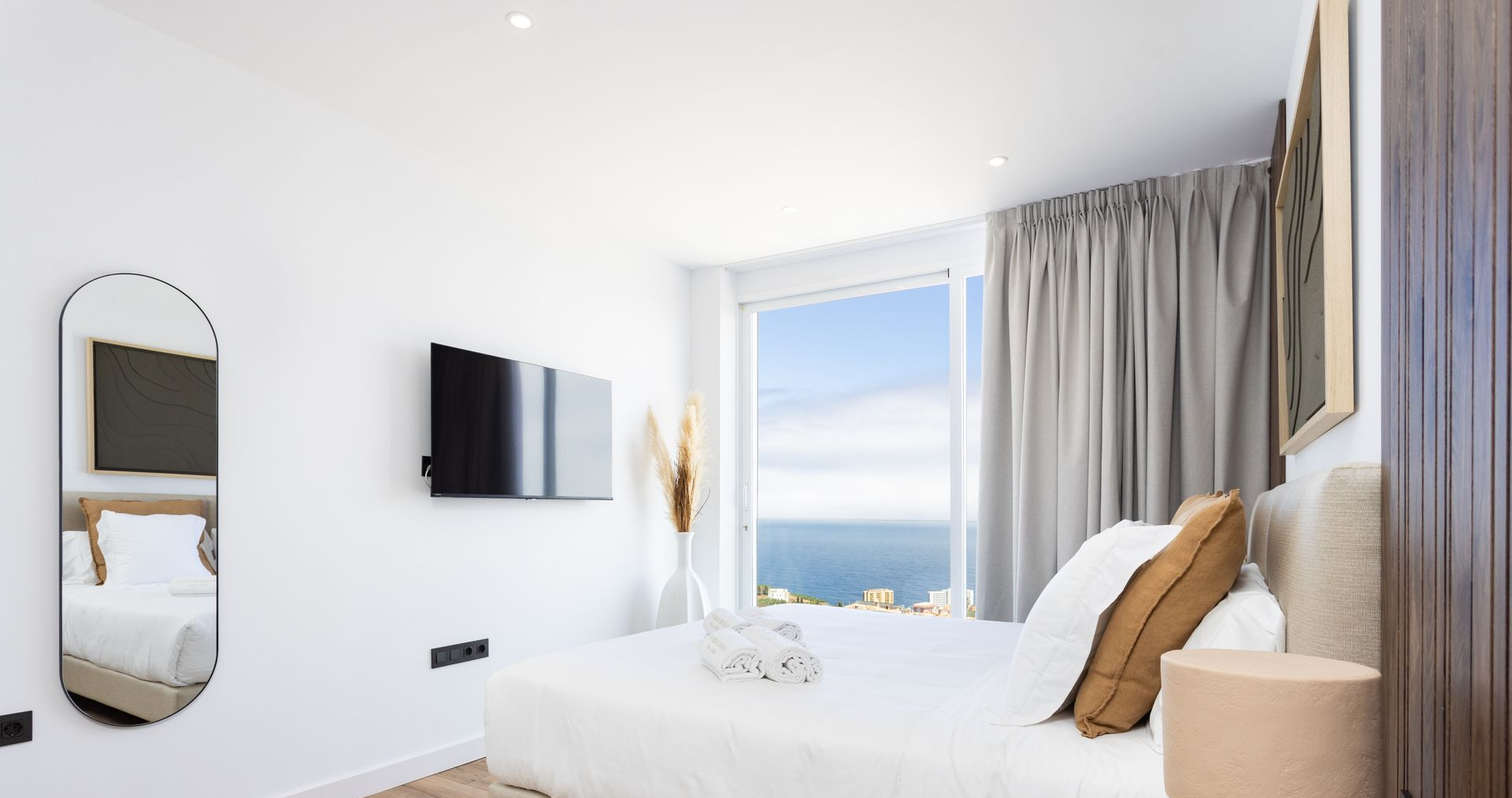 Habitación luminosa y minimalista con cama blanca, televisor montado en la pared, espejo y ventana con vistas al mar.