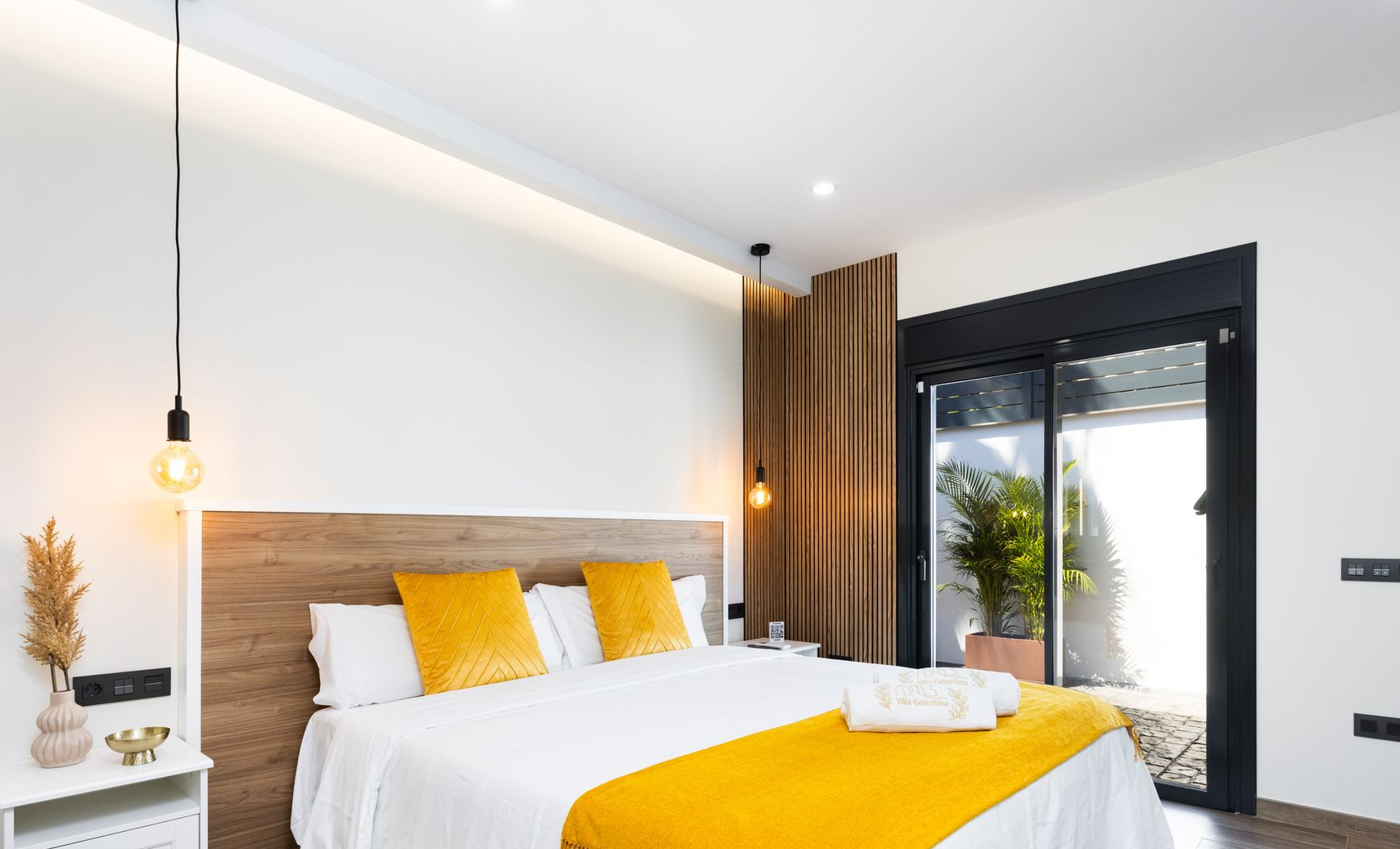 Dormitorio moderno con ropa de cama blanca, detalles en color mostaza, lámparas colgantes y una puerta de cristal que da a un patio.