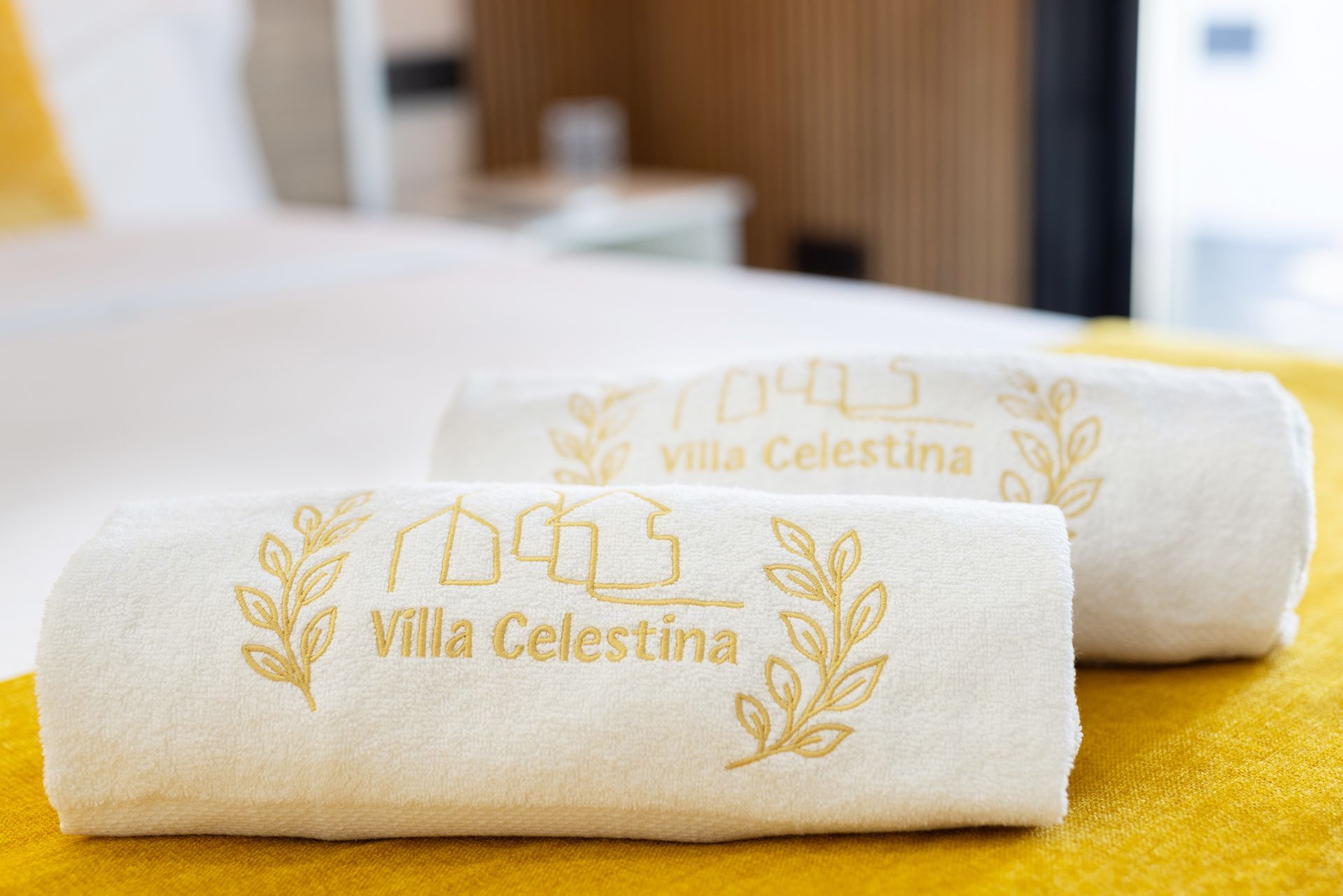 Dos toallas blancas enrolladas con bordado dorado “Villa Celestina” sobre una cama con una manta amarilla.
