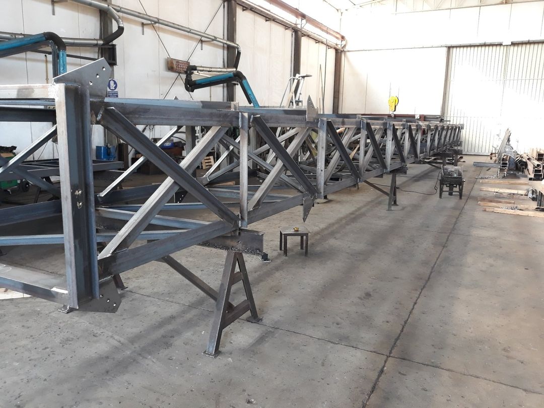 Lunga struttura reticolare in acciaio in fase di costruzione, sorretta da cavalletti metallici all'interno di un'officina industriale.