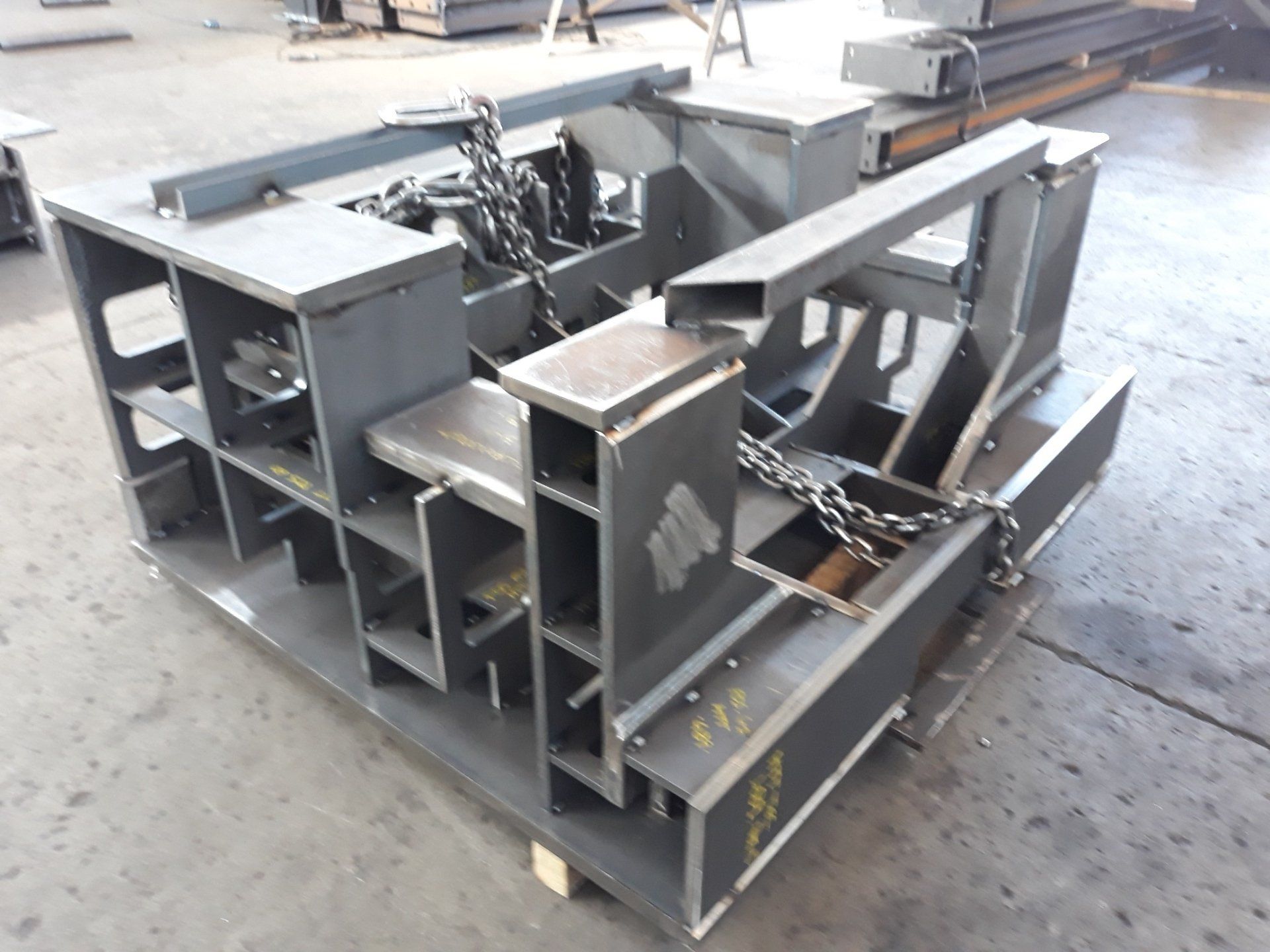 Una grande struttura industriale con telaio in acciaio, componenti saldati e catene metalliche, poggiata su un pavimento di cemento di una fabbrica.