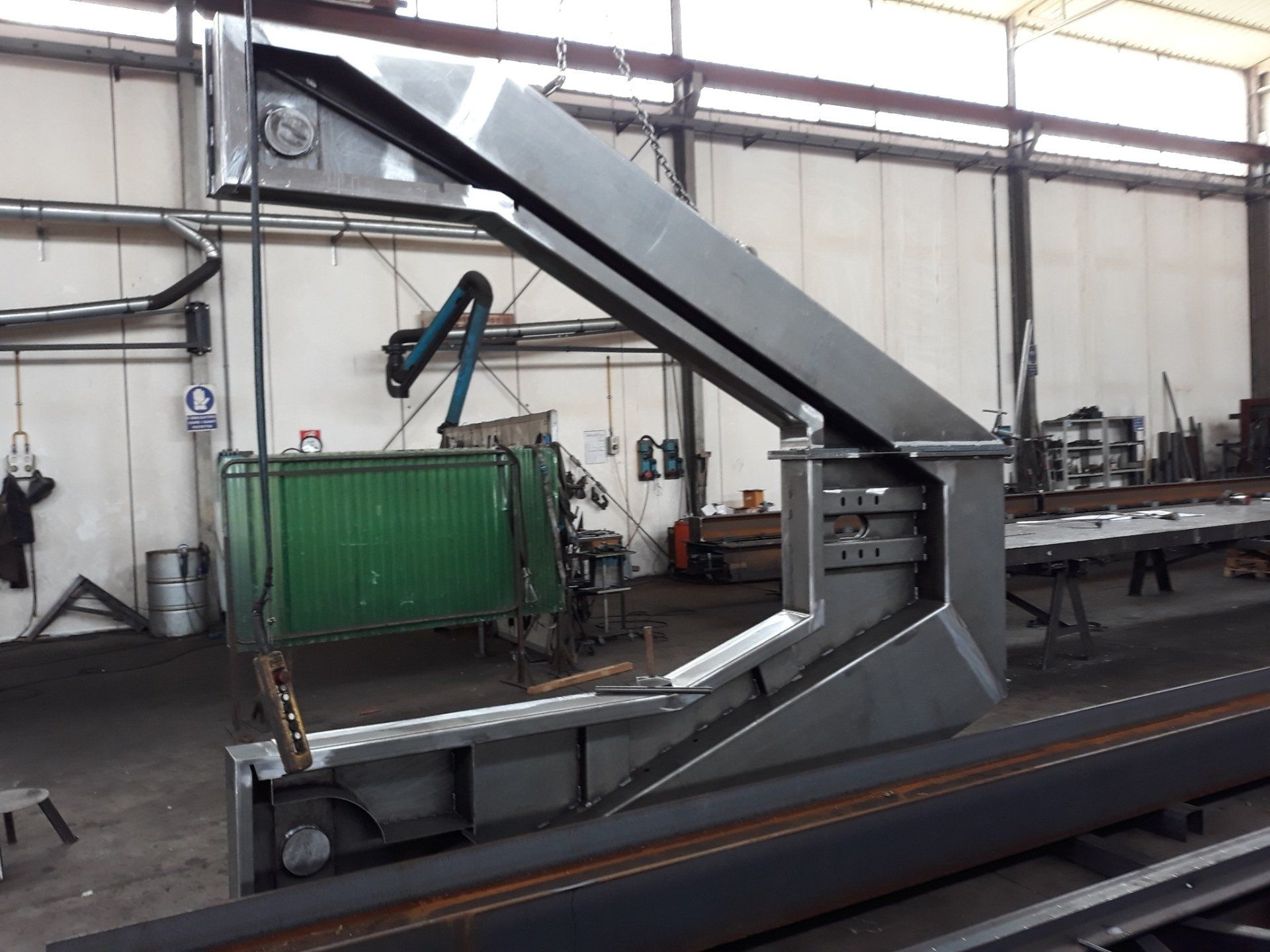 Una grande struttura angolare in acciaio grigio sospesa tramite un cavo all'interno di un'officina industriale, con macchinari sullo sfondo.