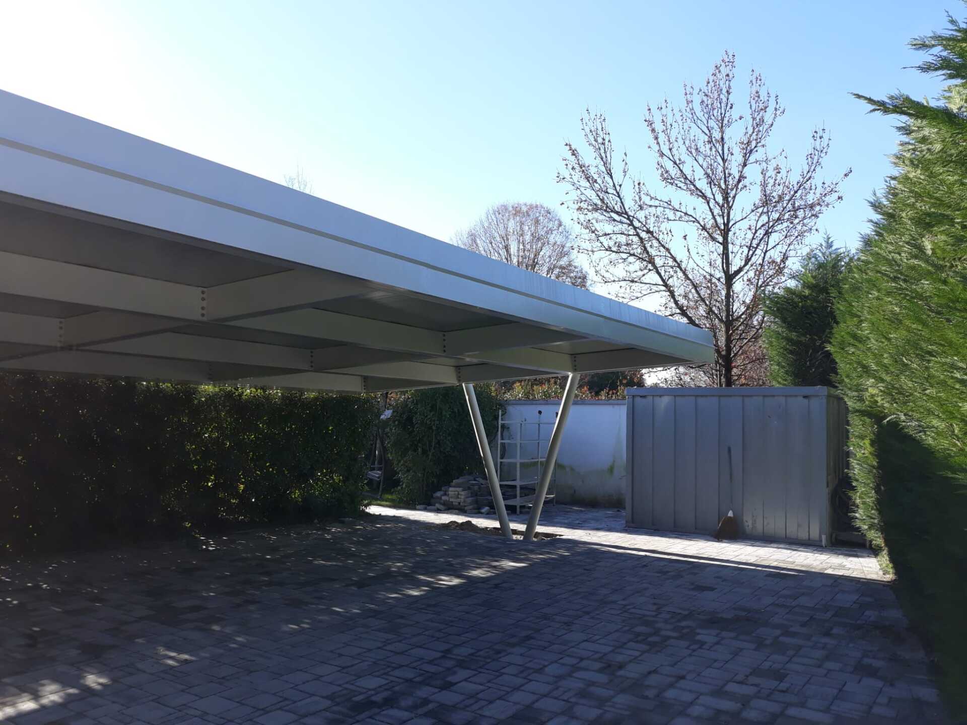 In un cortile residenziale, una moderna tettoia metallica con tetto piano e trave di supporto a forma di V si erge sopra un vialetto pavimentato.