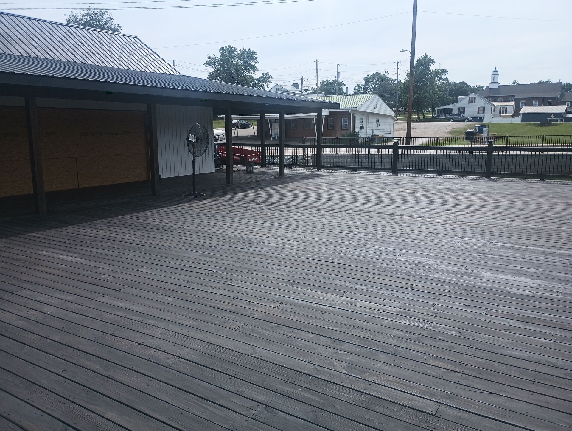 Empty wet wooden deck beside a covered building, with a fence and houses in the background.