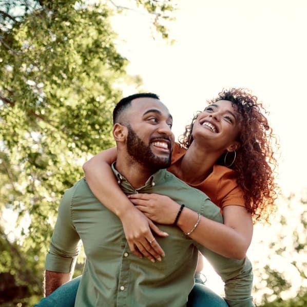 A smiling person gives another person a piggyback ride outdoors in a park setting.