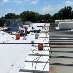 Roofing work on a flat roof under a blue sky; workers, tools, and materials are visible.