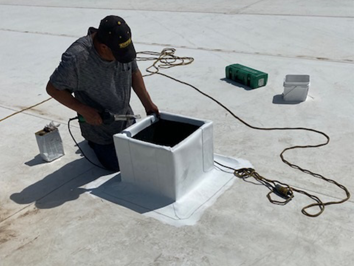 Person welding on a rooftop vent; white roofing material, tools, and a black vent opening are present.