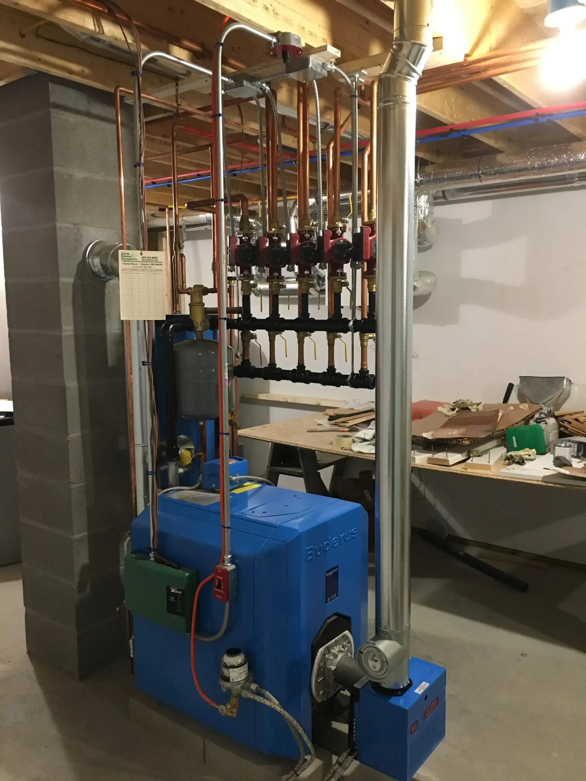 A blue boiler is sitting in a basement next to a pipe.