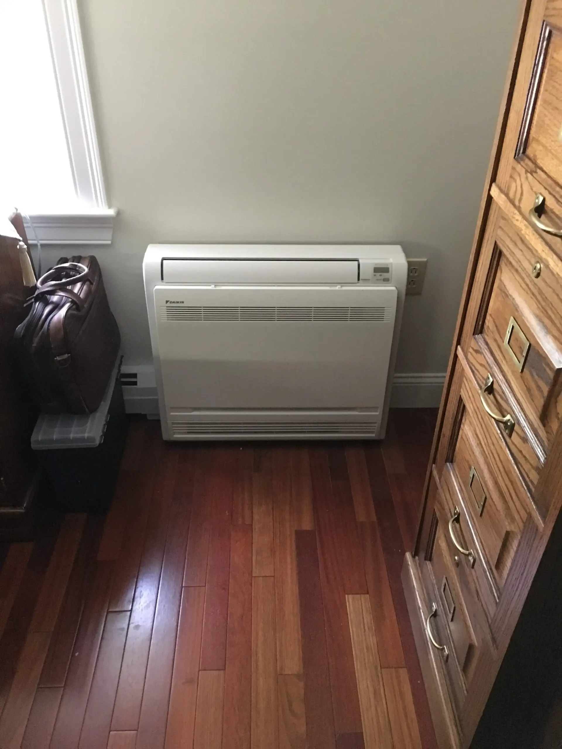A white air conditioner is sitting on a wooden floor in a room next to a window.