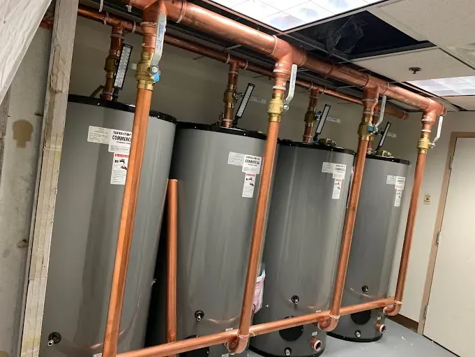 Four water heaters are lined up in a room with copper pipes.