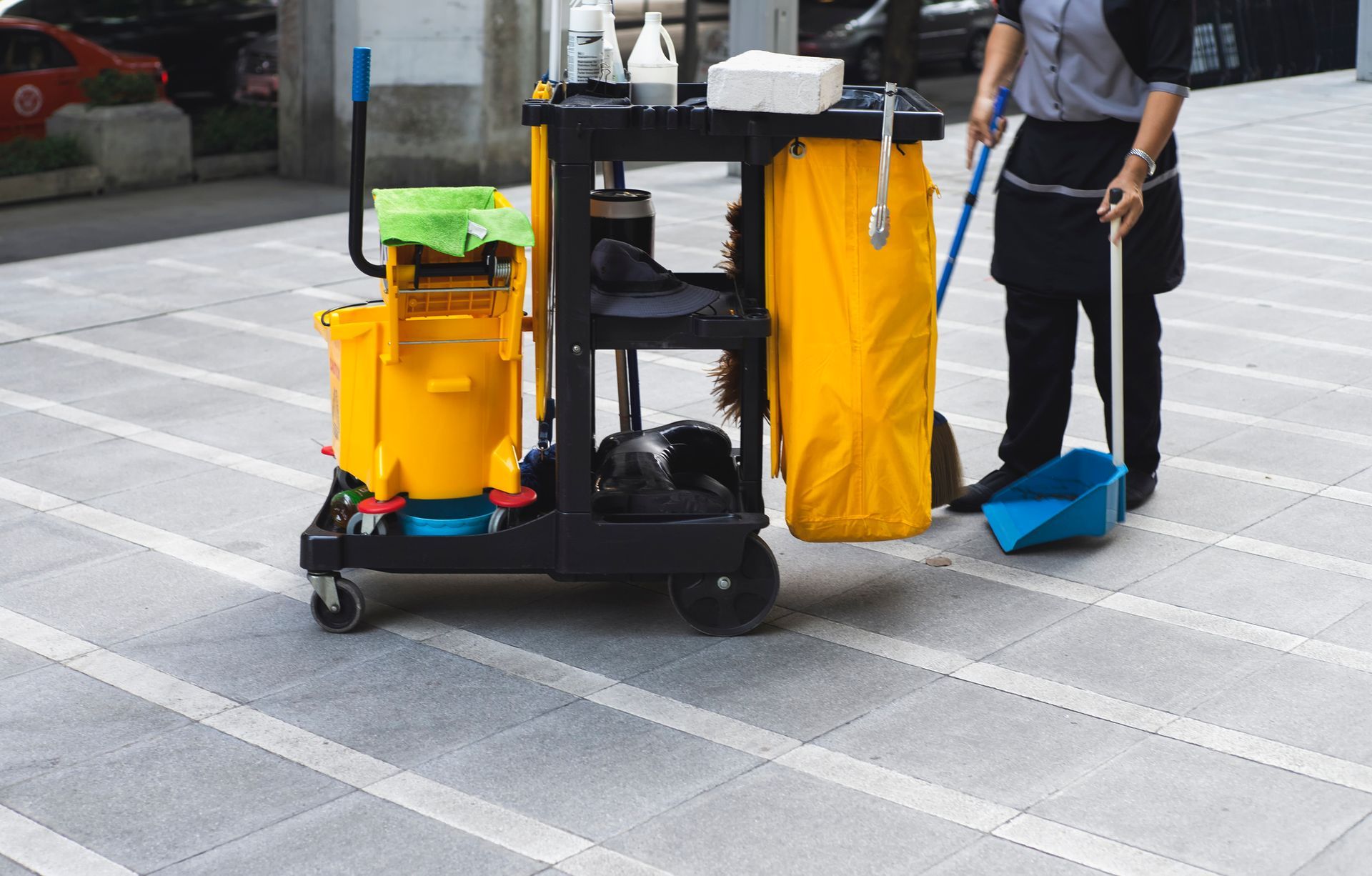 Janitor cart with yellow trash bags and cleaning supplies beside a person holding a broom on a sidewalk