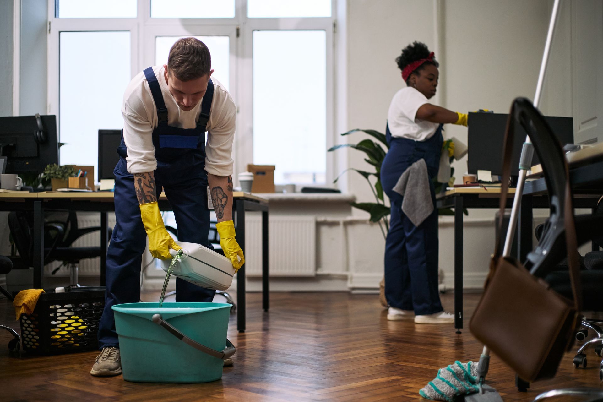 Two people cleaning an office floor with mops and buckets near desks and monitors