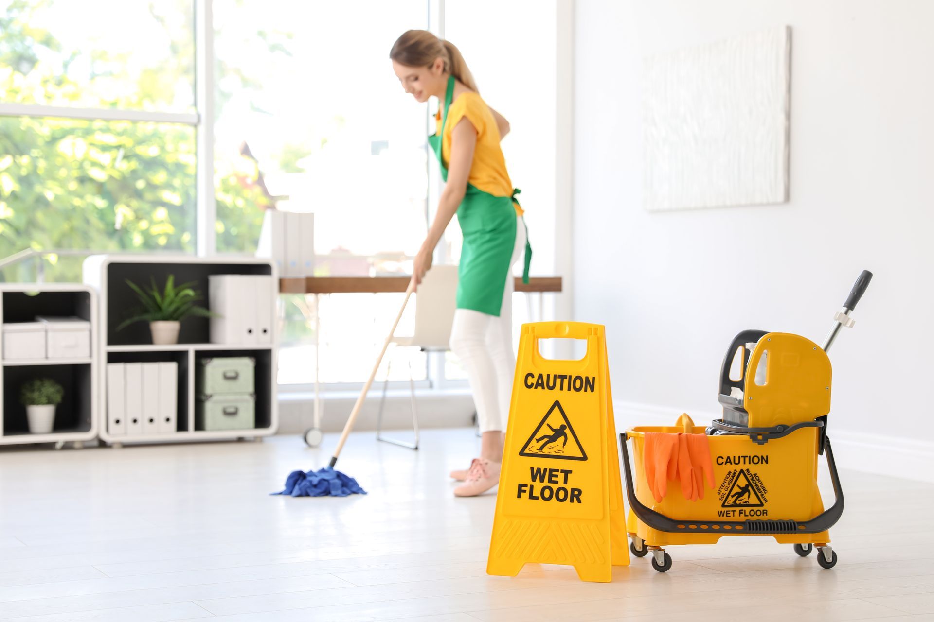 Woman mopping a bright room beside yellow caution floor signs and a cleaning cart