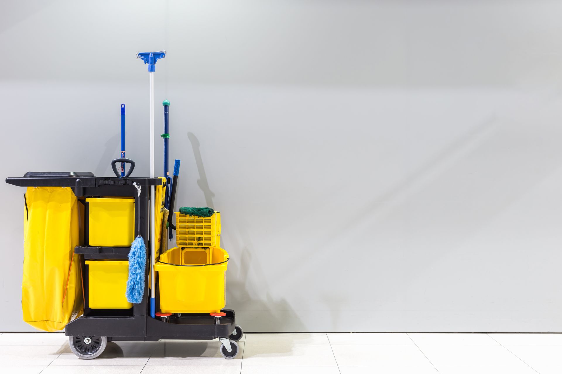 Yellow janitorial cleaning cart with mop, broom, bucket, and supplies on a white floor against a wall