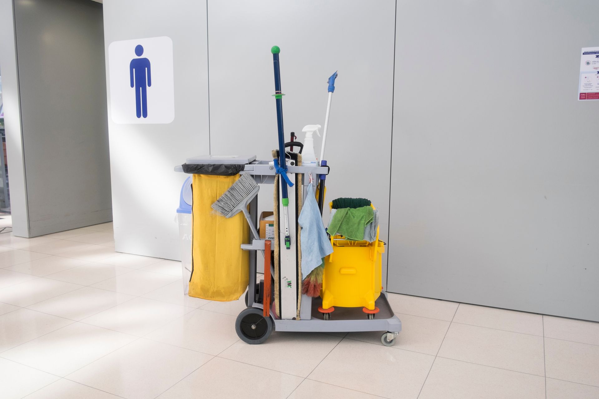Cleaning cart with mops and yellow bins in a hallway near a restroom sign