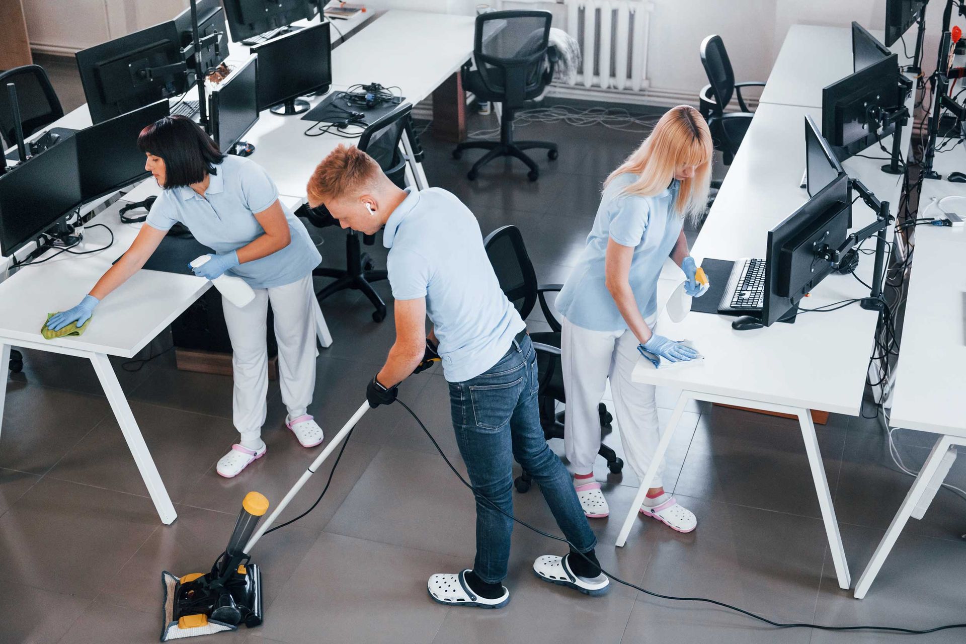 A group of workers is performing office cleaning solutions in a modern office during the day. Cleaning crew vacuuming and wiping desks in a bright office with computers and chairs