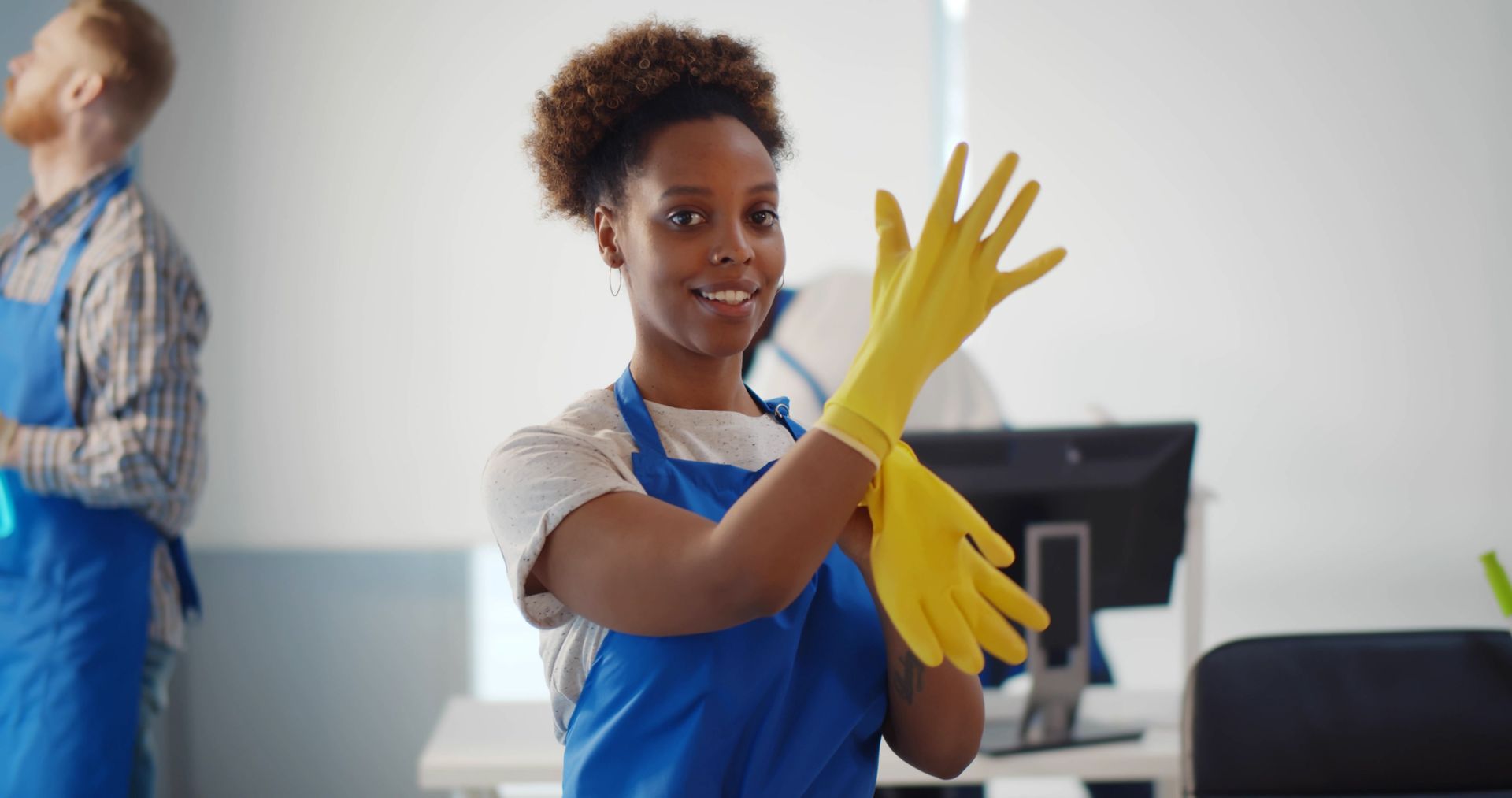 Worker in blue apron and yellow gloves, smiling in a cleaning setting while adjusting a glove.