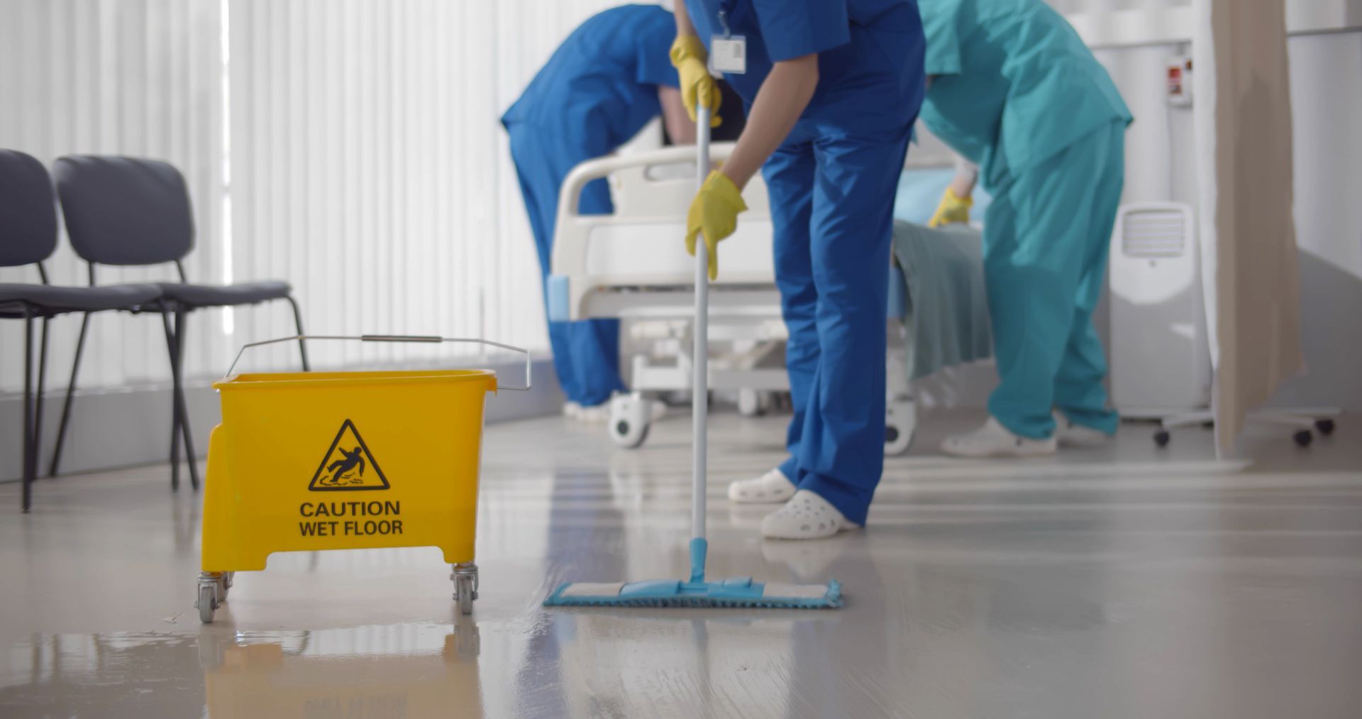 Hospital staff cleaning a floor beside a yellow wet floor warning sign and medical equipment.