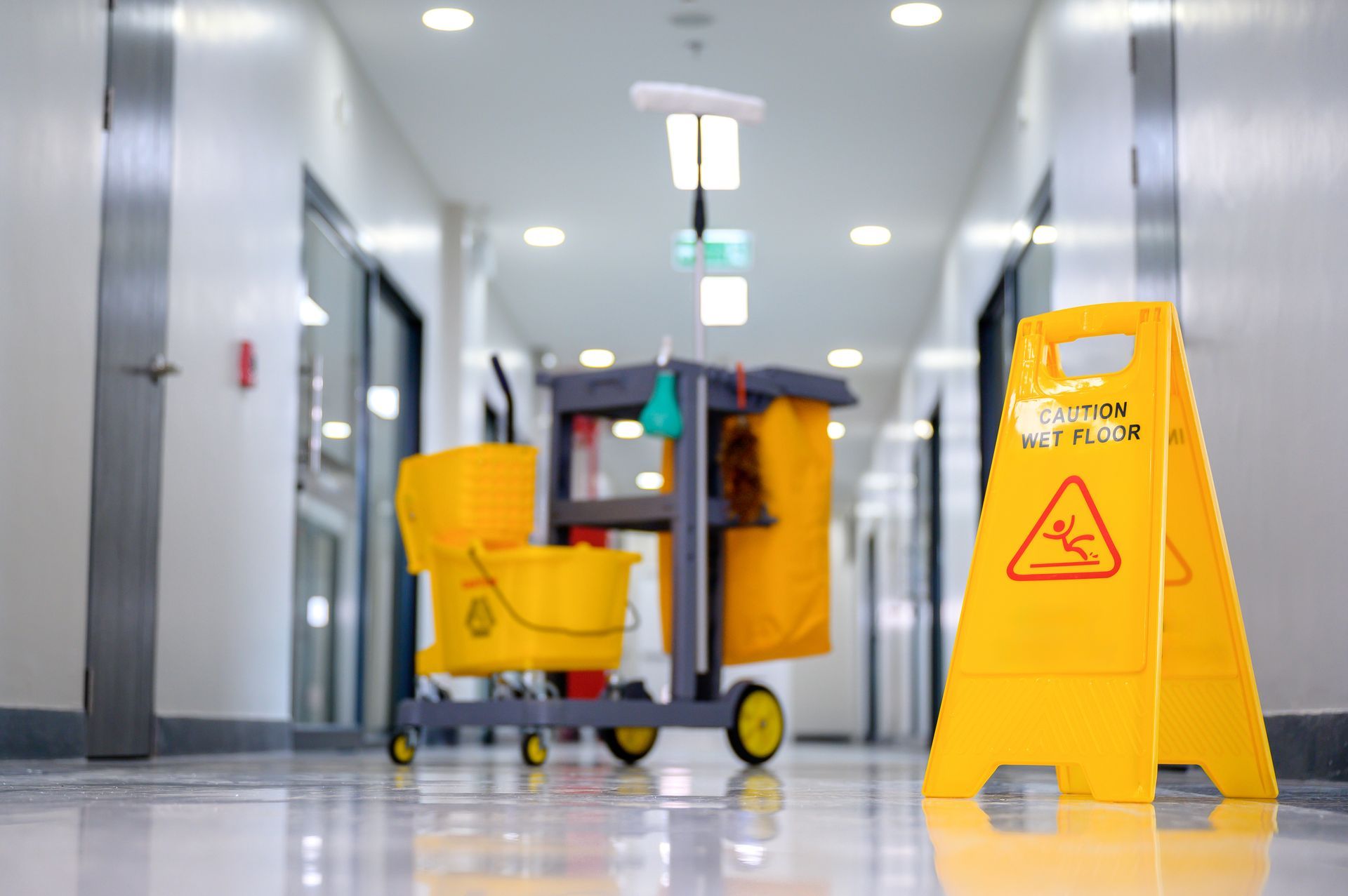 Yellow mop bucket and caution wet floor sign in a glossy hallway during cleaning.