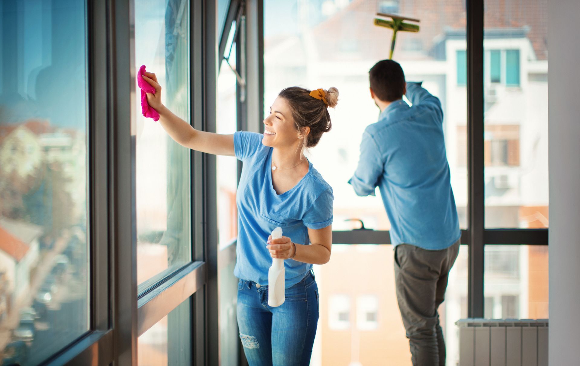 Two people cleaning large office windows, one wiping glass and one using a mop on the floor.