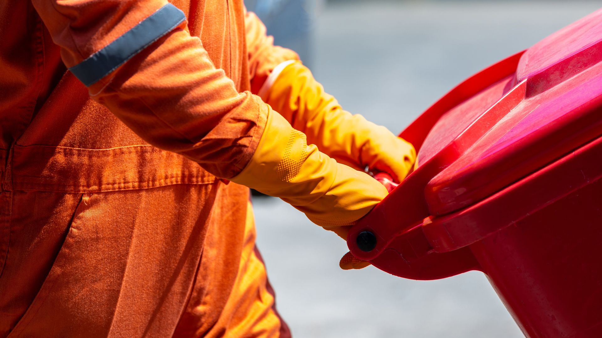Worker in orange gloves holding a red trash bin outdoors