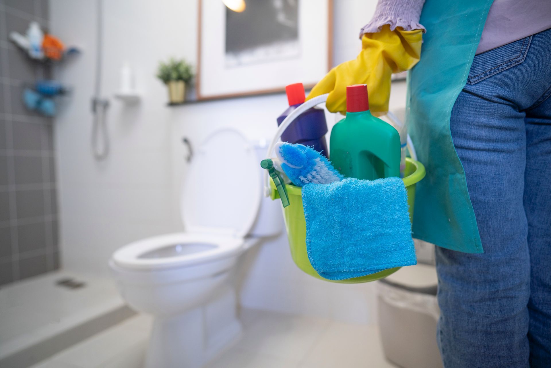 Person holding cleaning supplies in a bathroom, with a toilet and shower in the background.