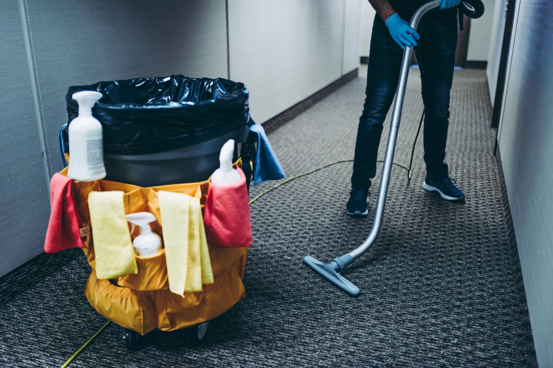 Custodian vacuuming carpet hallway beside a janitorial cart with cleaning supplies