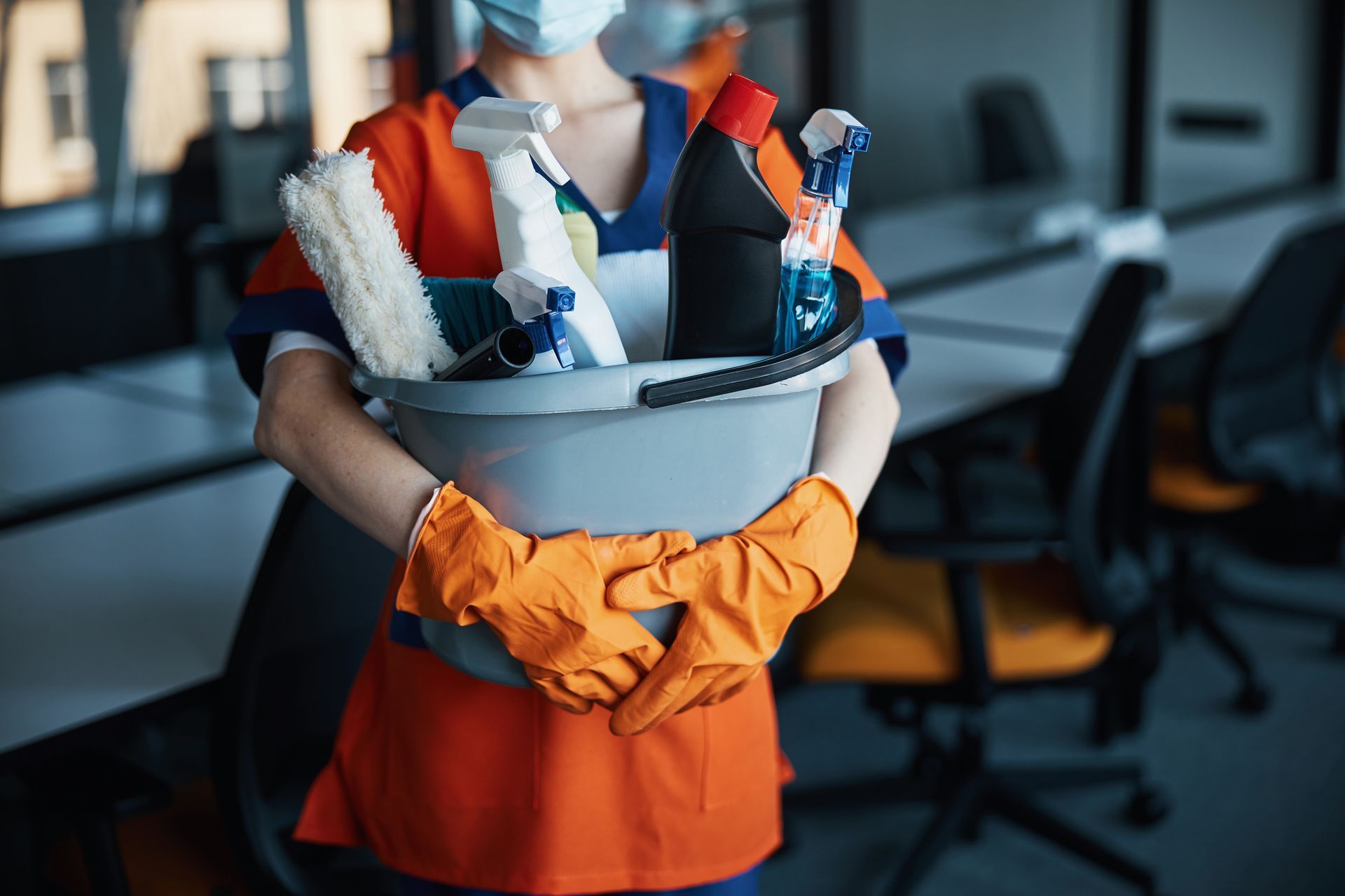 Person in orange gloves holding a cleaning bucket with spray bottles indoors
