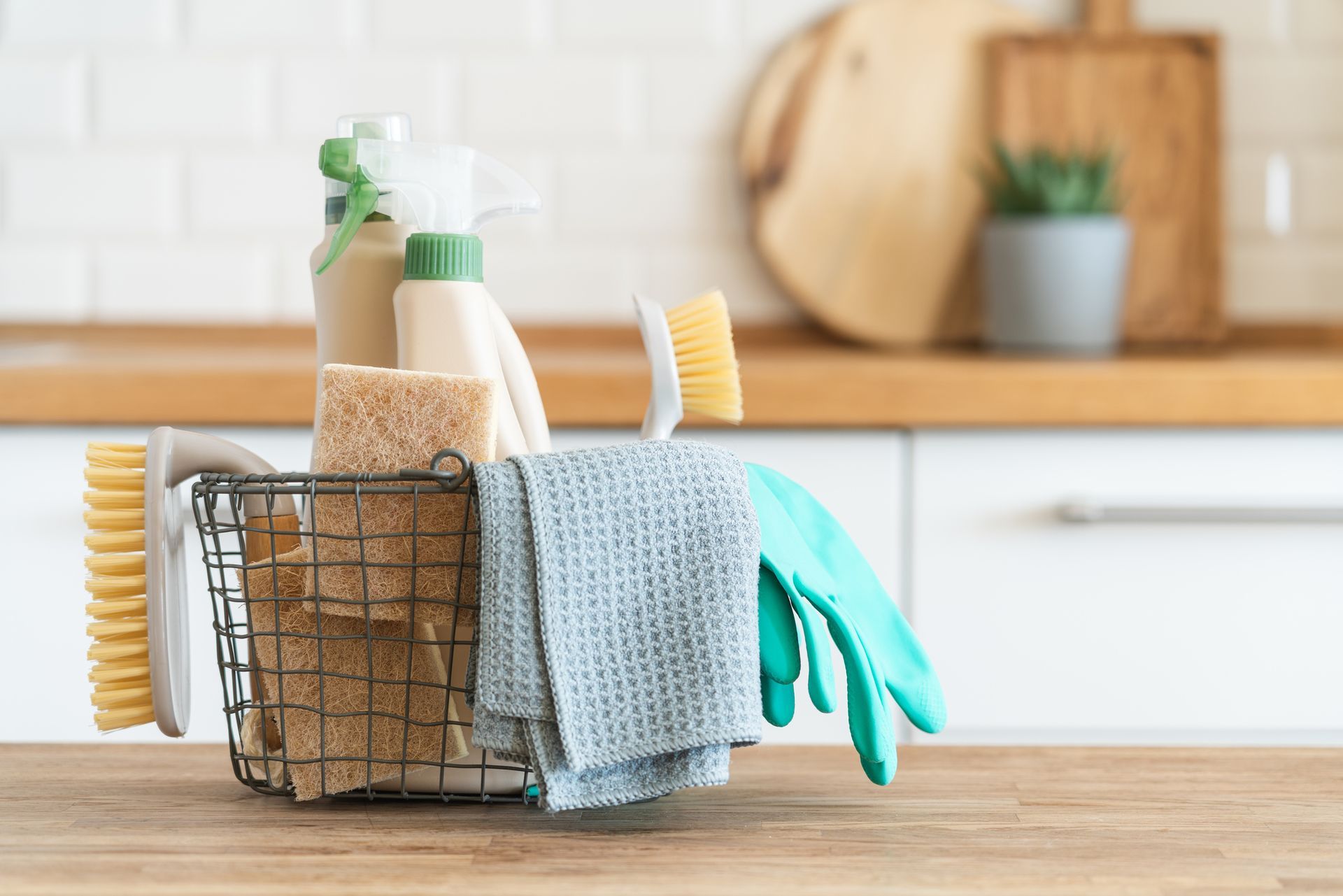 Cleaning supplies in a wire basket on a kitchen counter, with brushes, spray bottles, cloths, and gloves