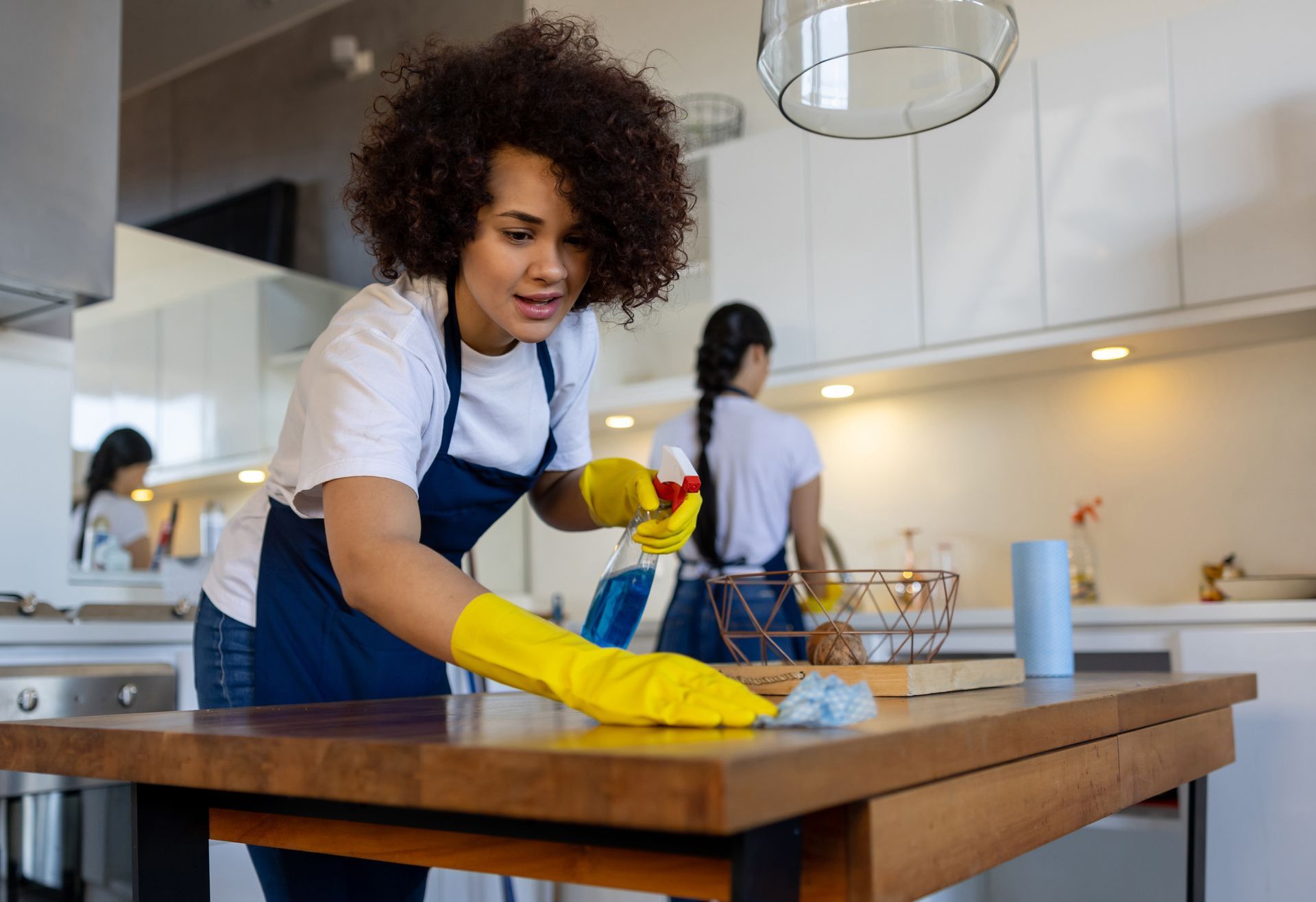 Janitorial cleaning service worker wiping wooden table with spray bottle in kitchen.