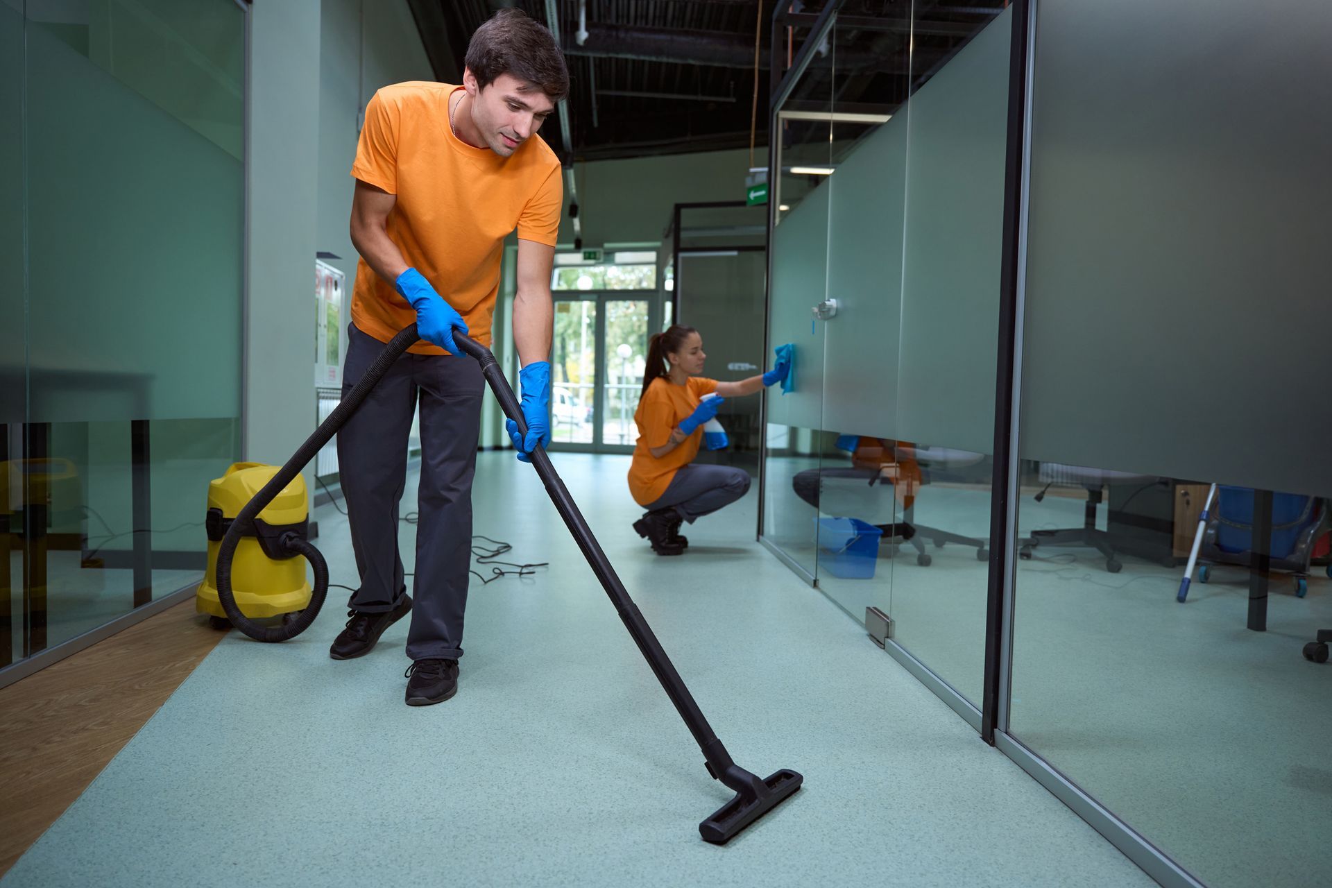Person vacuuming a glass-walled office hallway while another cleans in the background