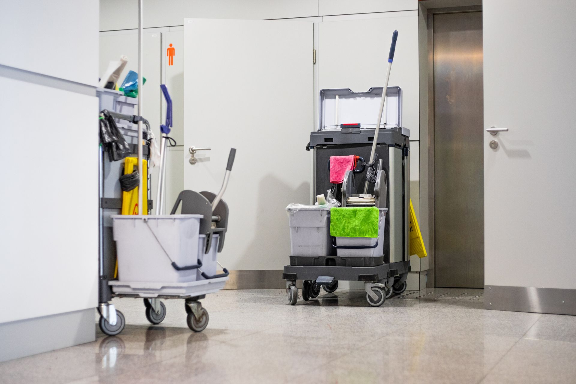 Two janitorial carts in a hospital hallway beside a stainless-steel elevator.