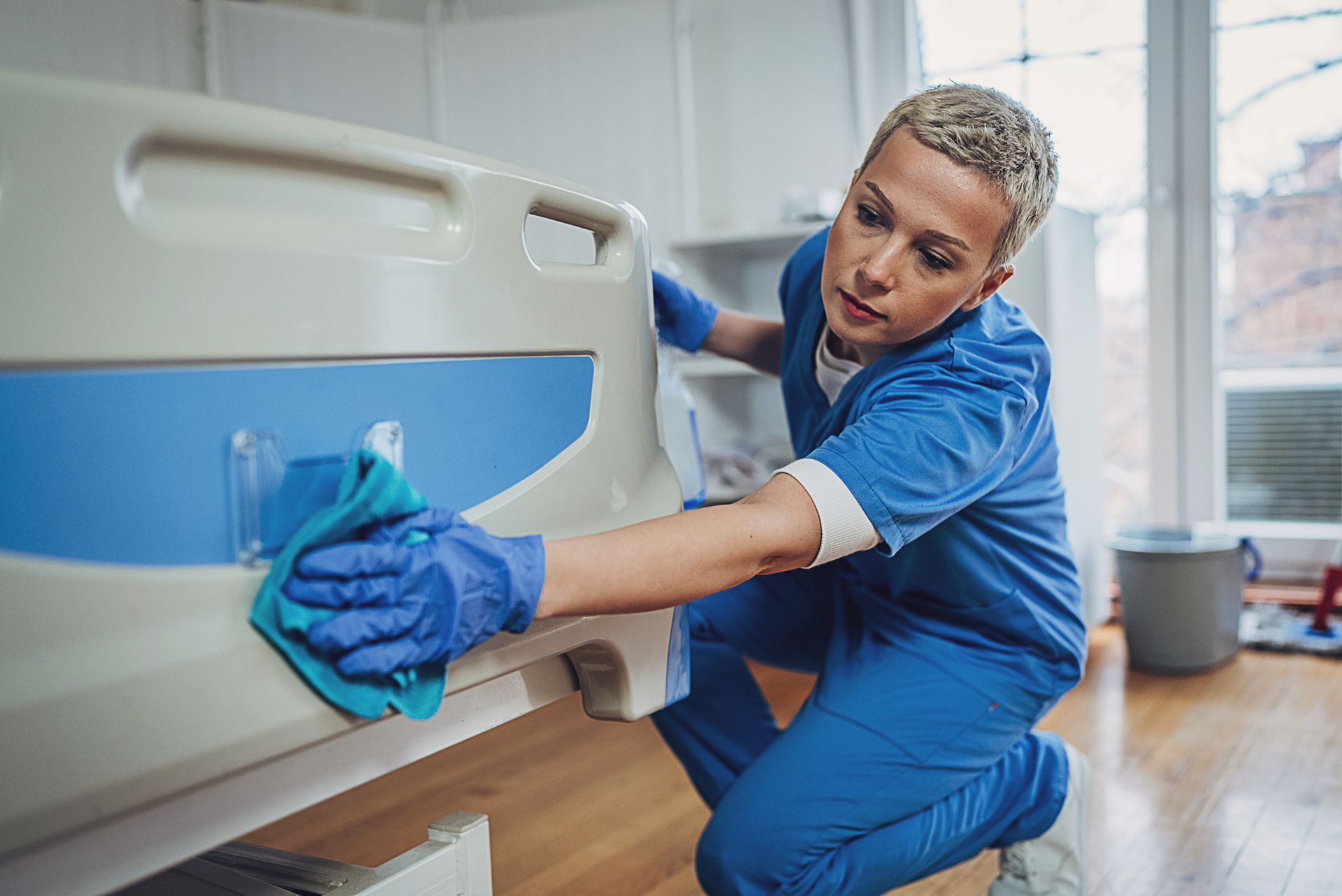 Person in blue scrubs cleaning a hospital bed with a cloth in a bright room