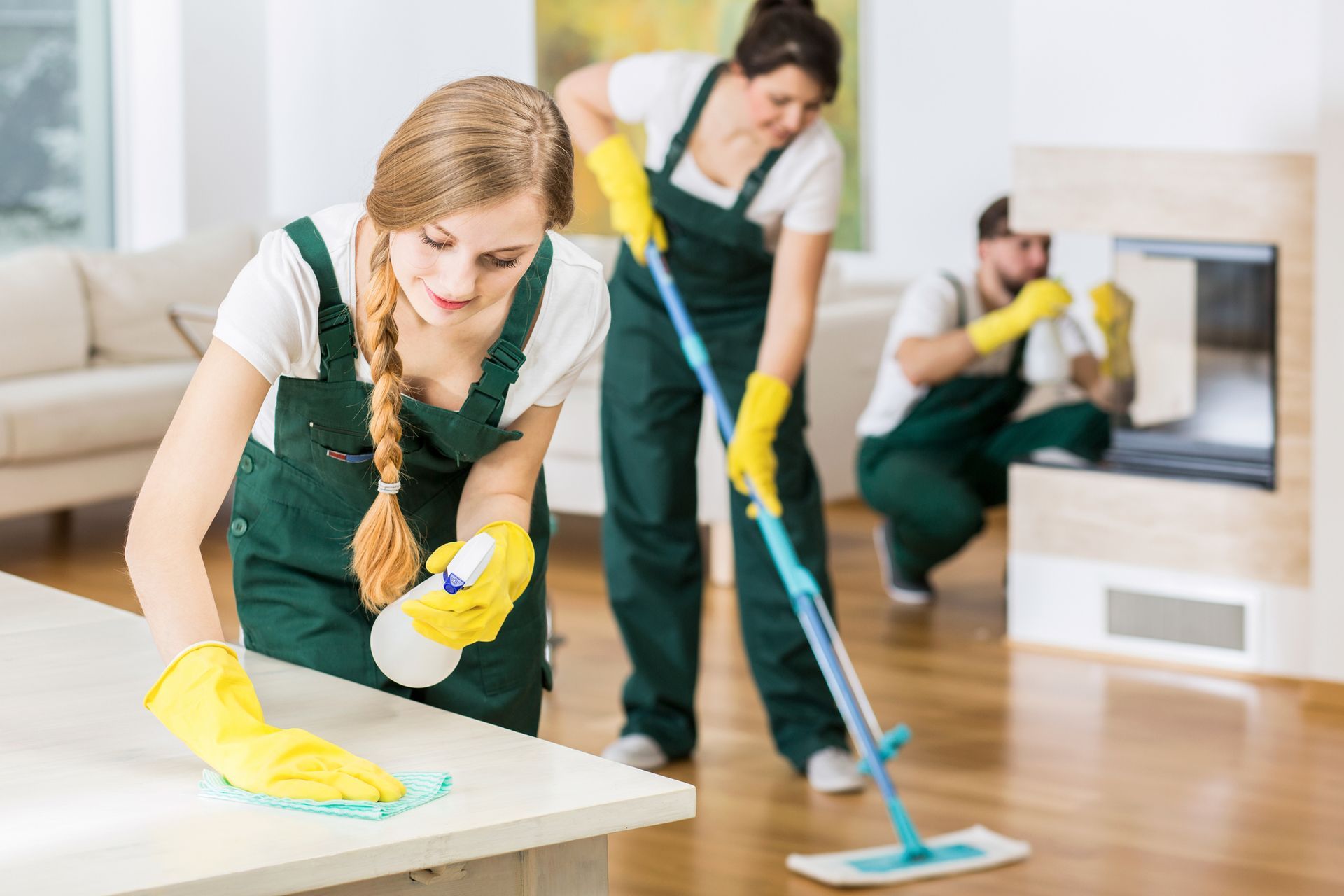 Three cleaners in green uniforms mopping and wiping a bright living room with a fireplace.
