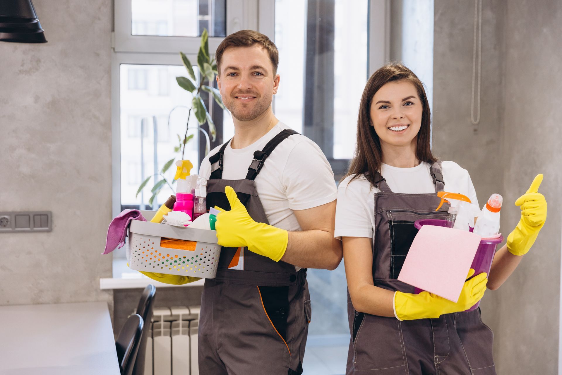 Two cleaners in yellow gloves holding cleaning supplies in a bright room