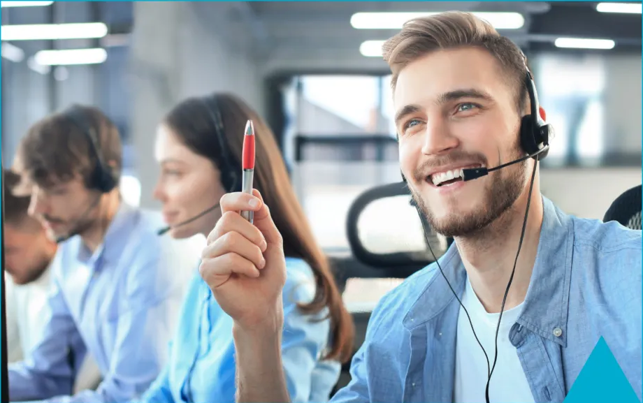 Agent de centre d'appels en chemise bleue souriant, casque sur les oreilles, stylo à la main, dans un bureau animé
