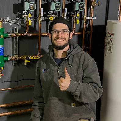 Smiling person in a beanie and glasses giving a thumbs-up in front of copper pipes and gauges.