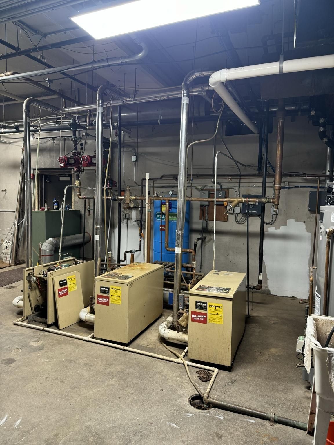 Industrial boiler room with three beige equipment units, pipes, valves, and overhead lights in a basement setting