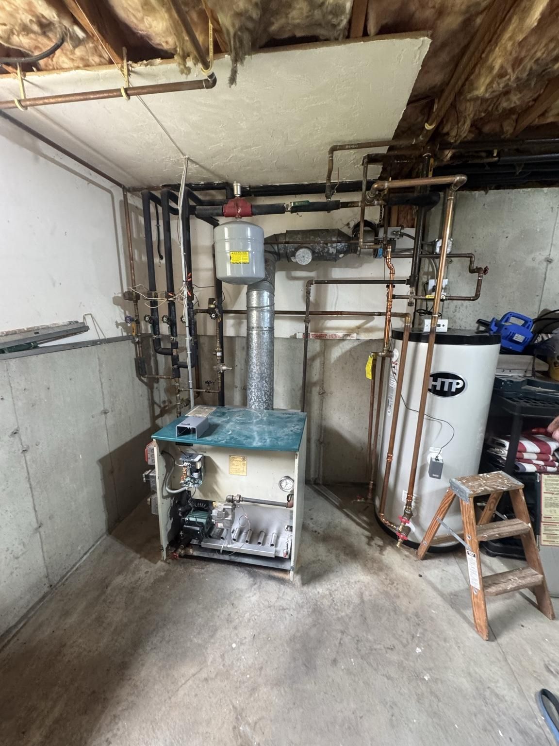 Basement utility room with furnace, water heater, exposed pipes, and a small ladder on a concrete floor