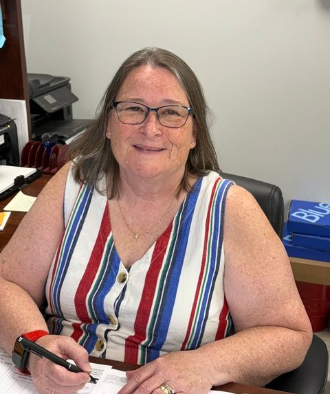 Woman seated at a desk, smiling and holding a pen, wearing a striped sleeveless top and glasses.
