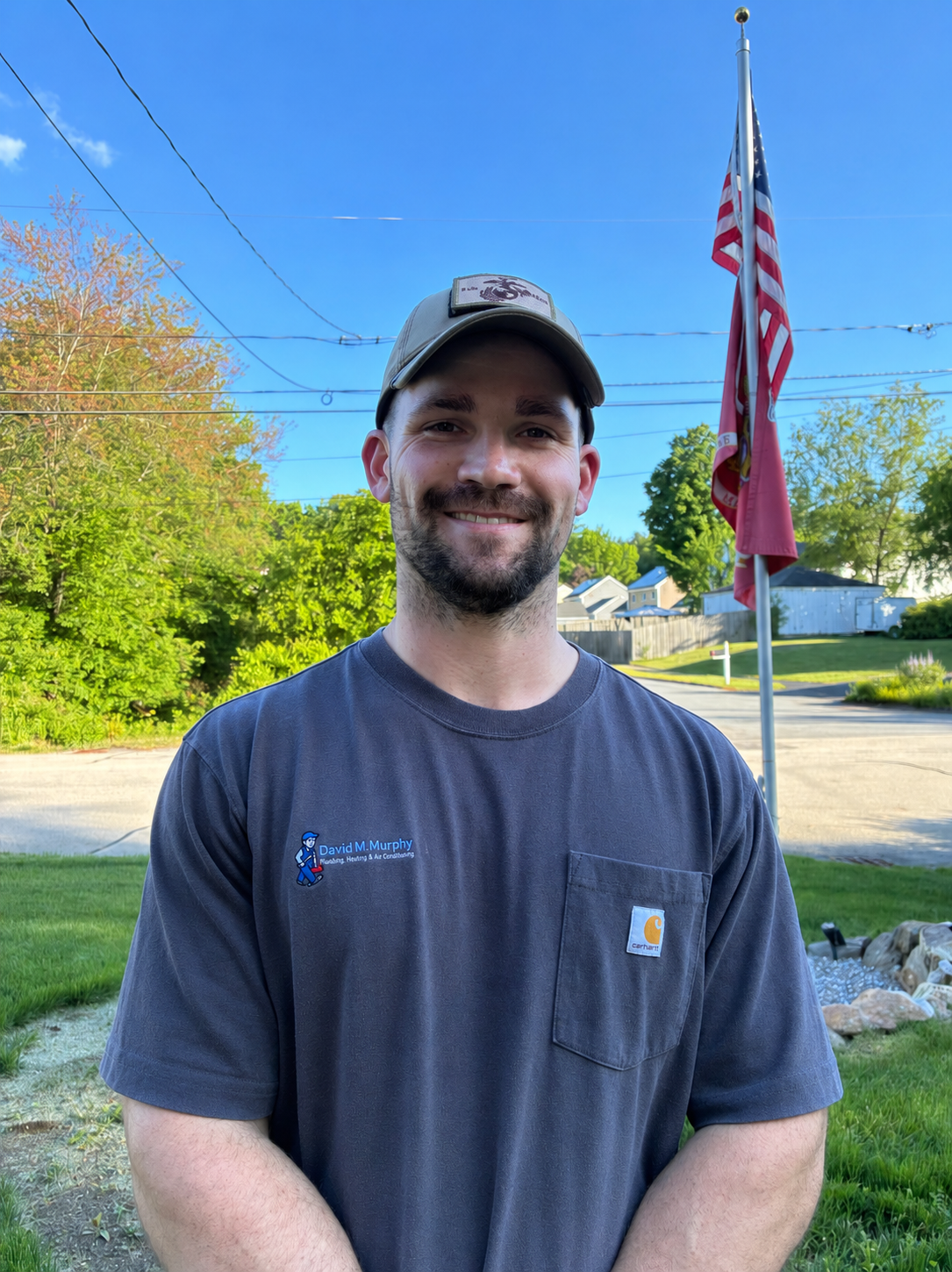 Smiling man in a cap and gray T-shirt standing outdoors with an American flag in the background