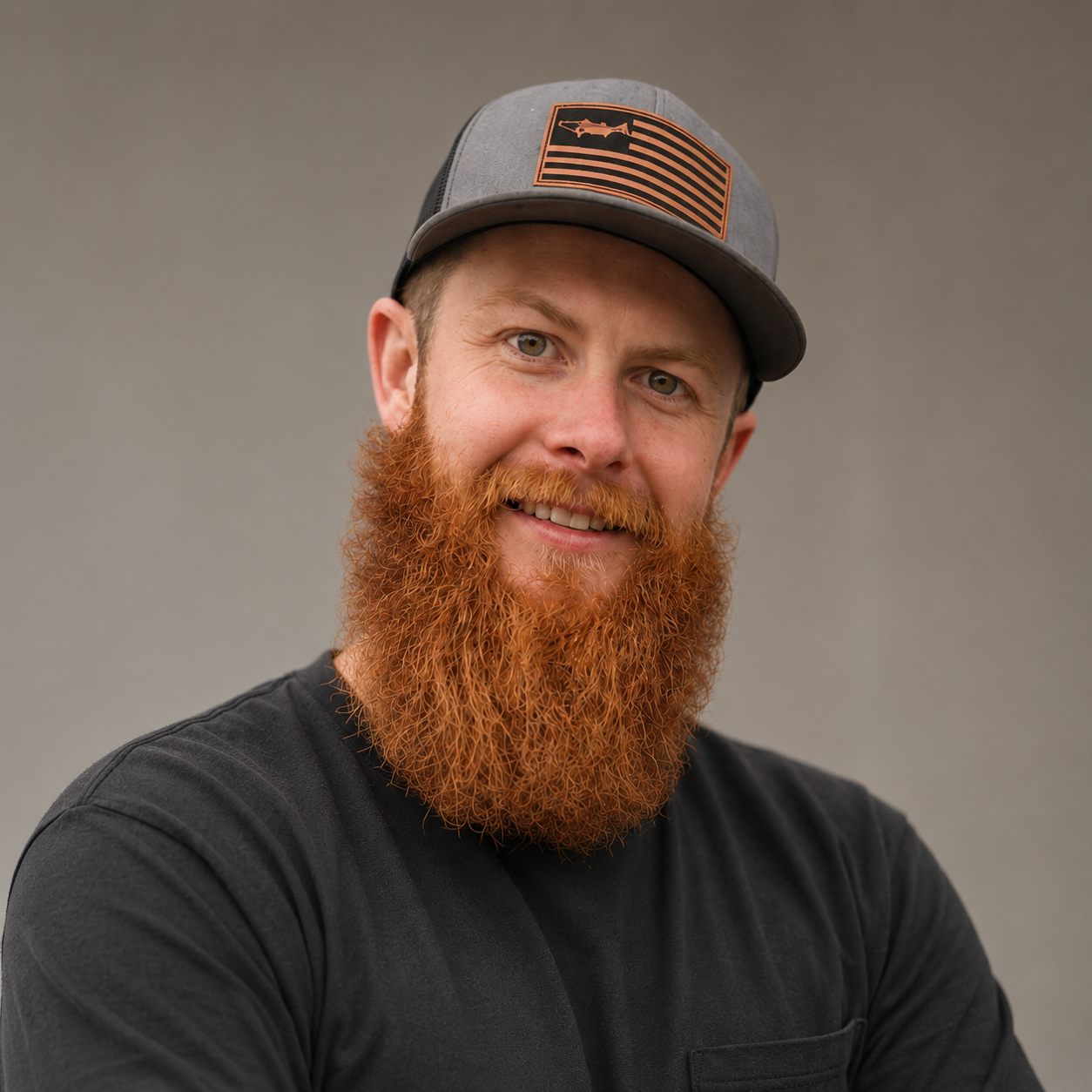 Portrait of a smiling man with a red beard wearing a gray cap and black shirt against a neutral background