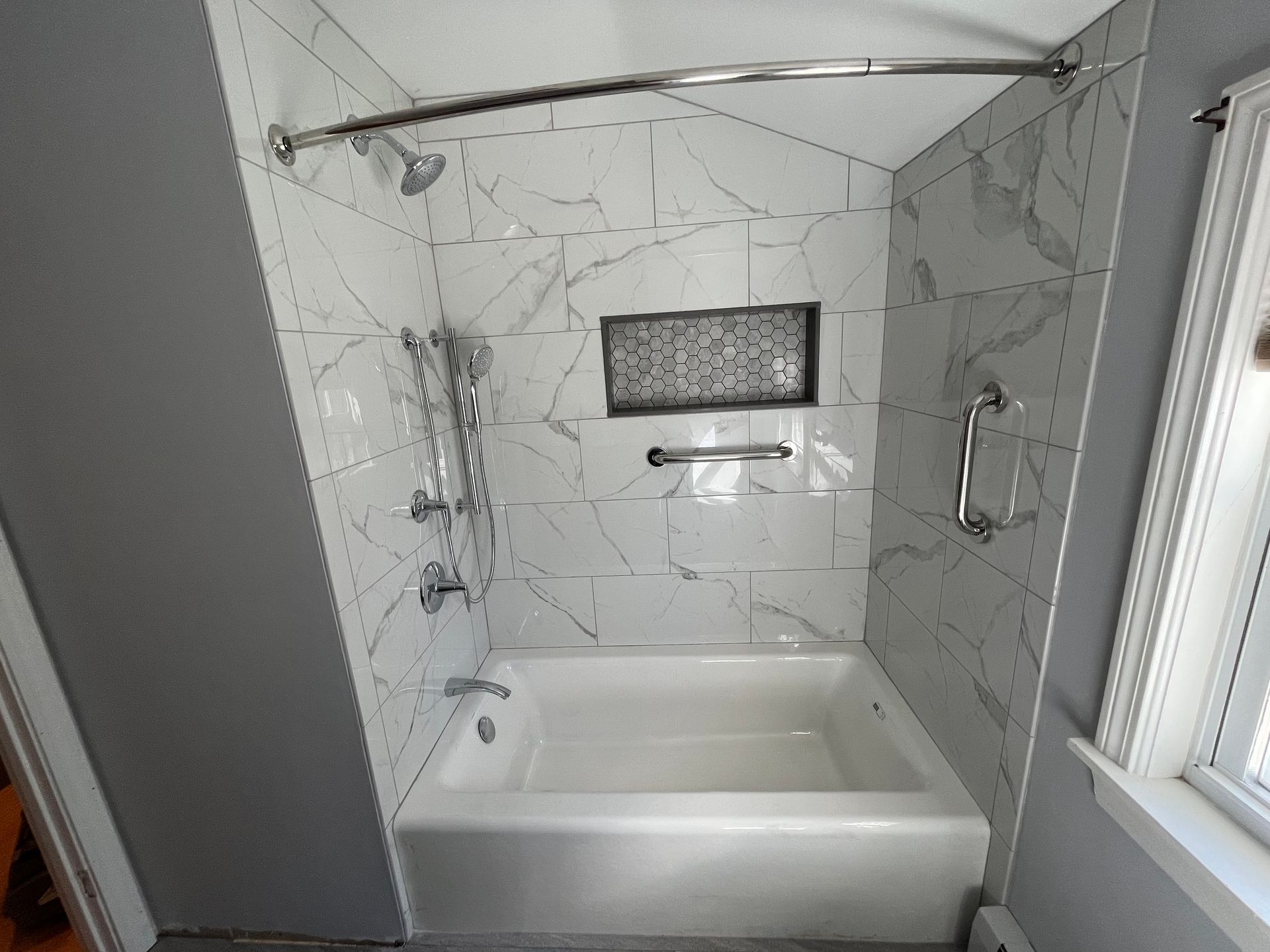 Modern tiled bathroom with a white tub, grab bars, handheld shower, and window.