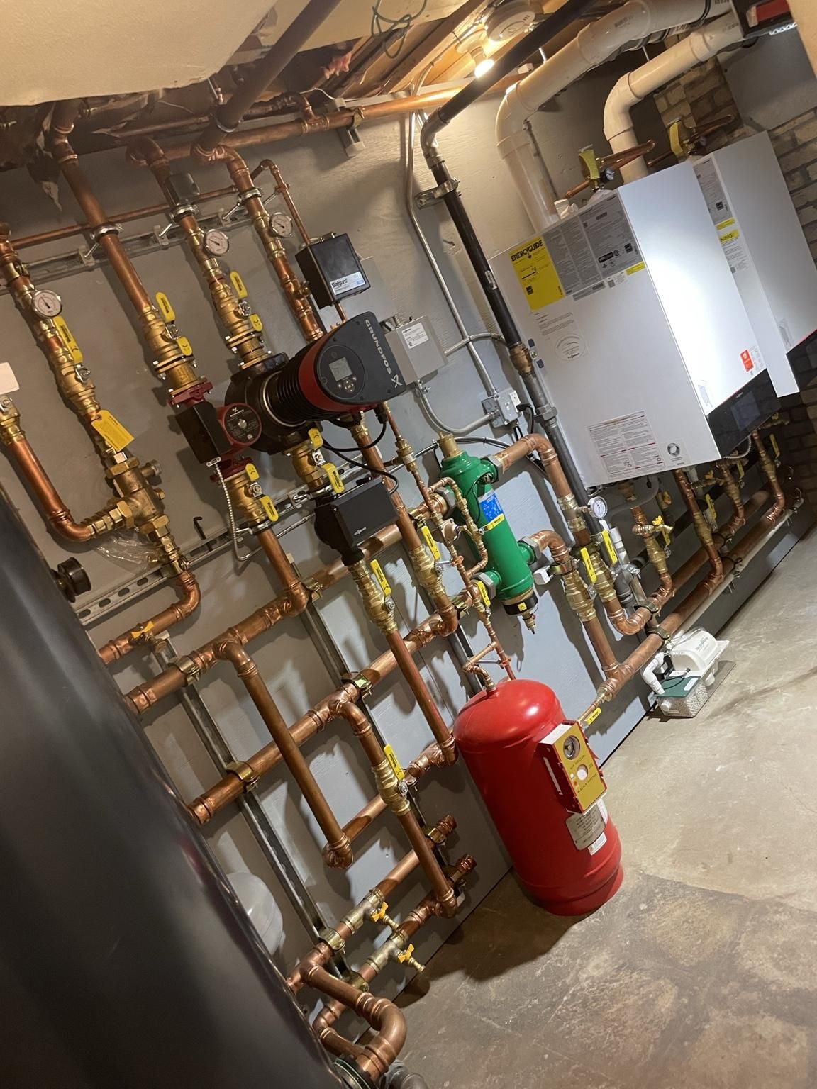 Basement utility room with copper pipes, gauges, white boiler units, and a red fire extinguisher.