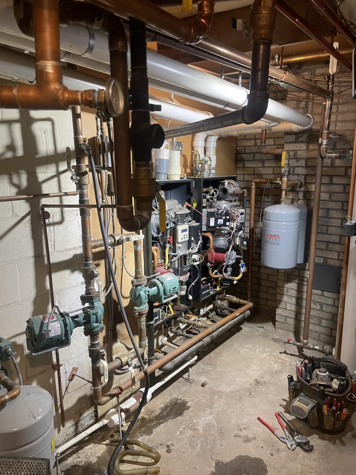 Basement mechanical room with copper pipes, pumps, boiler equipment, and storage tank in a utility space
