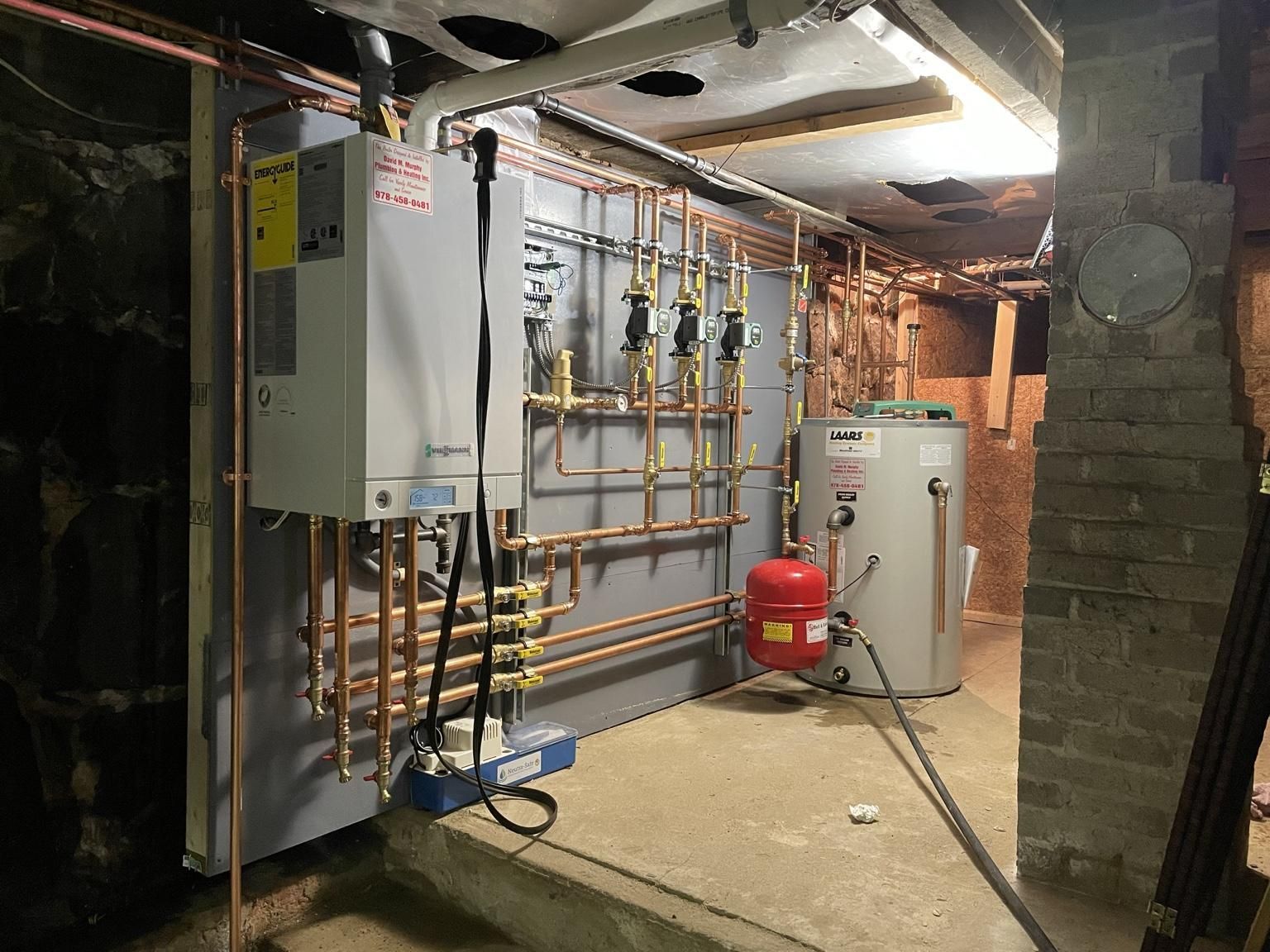 Basement utility room with plumbing pipes, control panel, water heater, and red expansion tank on a concrete floor